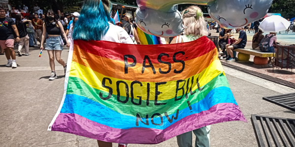 Photo of an LGBTQ couple wearing the pride flag with a call to pass SOGIE Bill on their backs